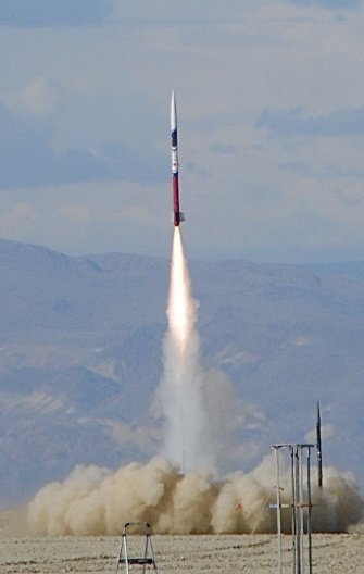 A rocket lifting off from the desert launch site against a backdrop of mountains