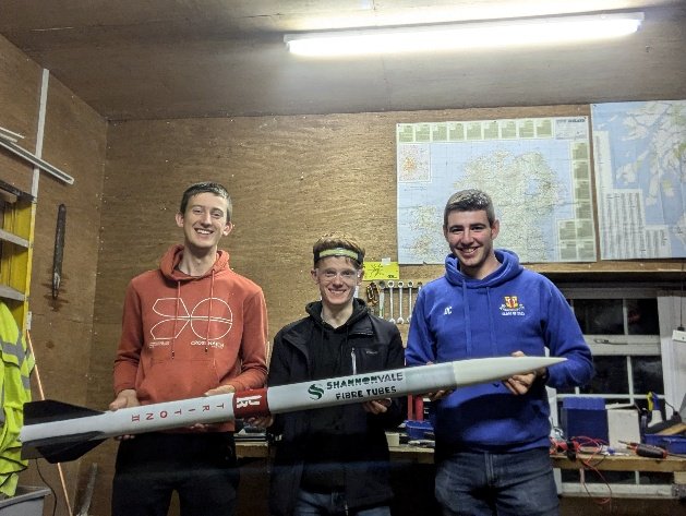 Three team members holding the rebuilt Triton II rocket in the workshop
